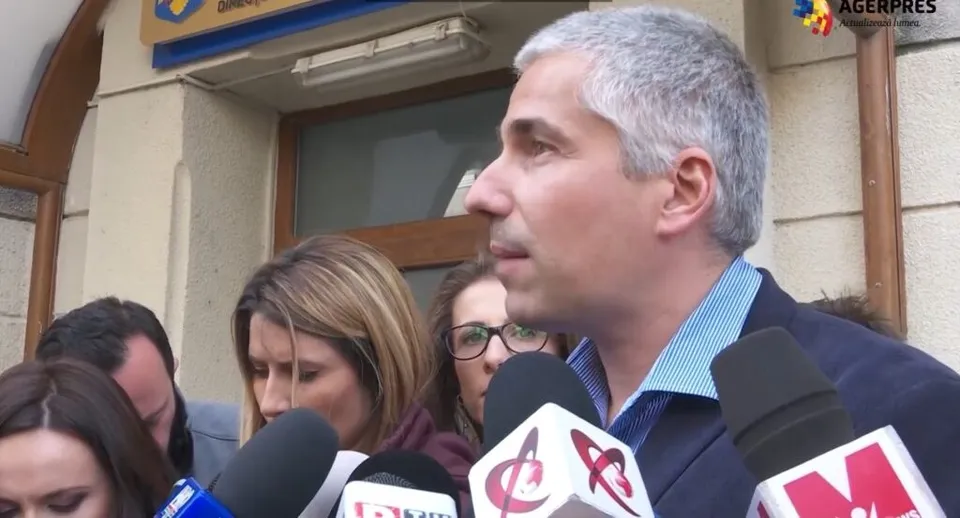Un bărbat cu părul scurt și cămașă albastră este înconjurat de microfoane și persoane în plan secund în fața unei clădiri.