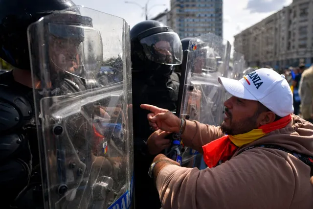 Un bărbat cu șapcă albă și eșarfă colorată arată spre polițiști care poartă echipament de protecție și scuturi transparente.