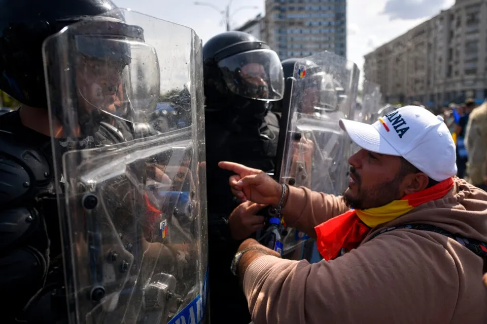 Un bărbat cu șapcă albă și eșarfă colorată arată spre polițiști care poartă echipament de protecție și scuturi transparente.