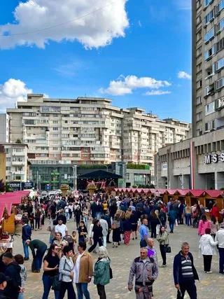 O mulțime de oameni merge pe o stradă pietonală între clădiri înalte și tarabe de vânzare sub un cer cu nori albi.