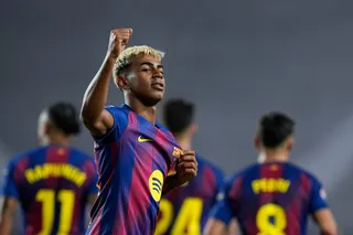 Un jucător de fotbal cu părul deschis la culoare ridică un pumn în sus, purtând un echipament sportiv albastru și roșu, în fața altor jucători.