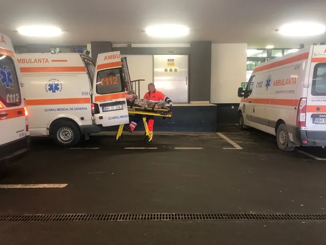 Două ambulanțe albe cu marcaje portocalii sunt parcate într-un spațiu acoperit, lângă o targă de transport cu o persoană în poziție orizontală.