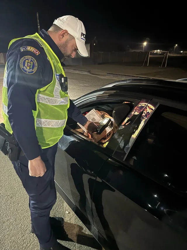 Un polițist cu vestă reflectorizantă și șapcă stă lângă o mașină neagră și privește spre interiorul acesteia pe timp de noapte.