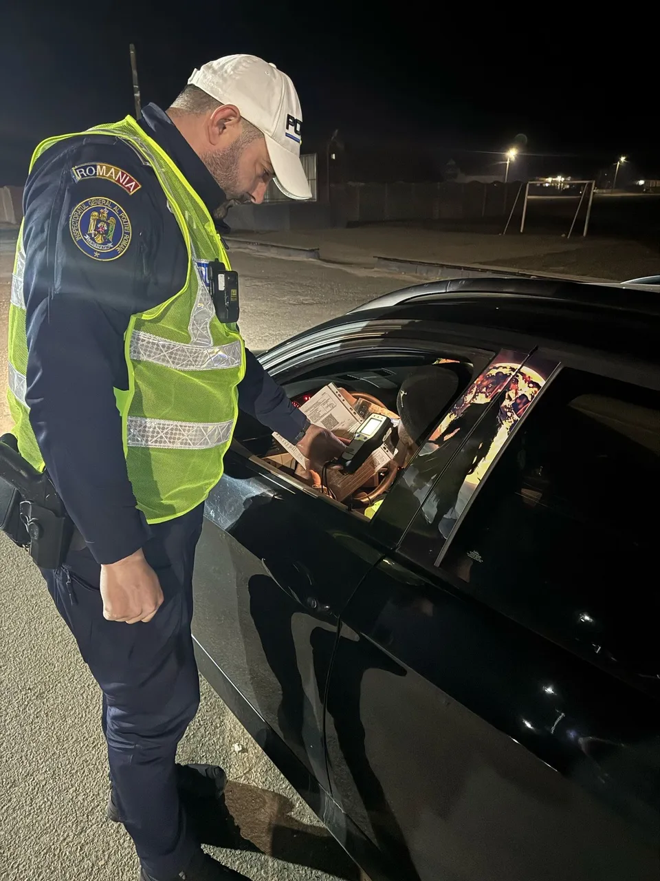 Un polițist cu vestă reflectorizantă și șapcă stă lângă o mașină neagră și privește spre interiorul acesteia pe timp de noapte.
