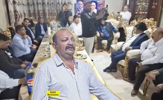 Un bărbat cu mustață și cămașă în dungi este suprapus în prim-plan, în timp ce un grup de persoane stă așezat în jurul unei mese într-o încăpere.