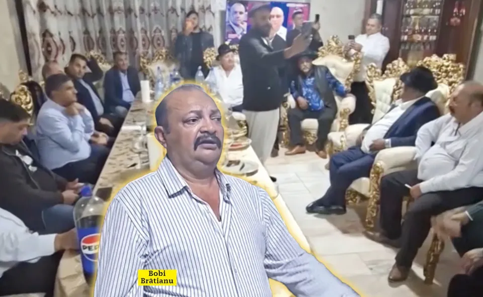 Un bărbat cu mustață și cămașă în dungi este suprapus în prim-plan, în timp ce un grup de persoane stă așezat în jurul unei mese într-o încăpere.