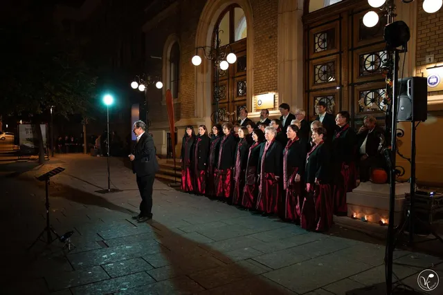 Un grup de persoane îmbrăcate în haine negre și roșii cântă în fața unei clădiri mari, sub lumina unui stâlp de iluminat nocturn.