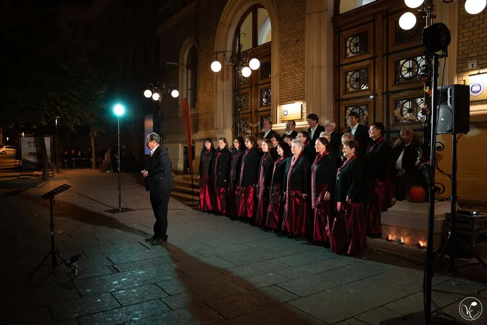 Un grup de persoane îmbrăcate în haine negre și roșii cântă în fața unei clădiri mari, sub lumina unui stâlp de iluminat nocturn.