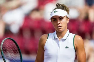 O femeie cu vizieră albă și tricou alb fără mâneci ține o rachetă de tenis în fața unui fundal colorat și neclar.