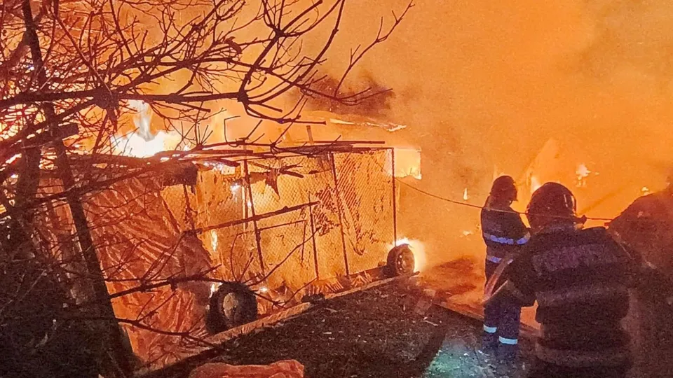 Două persoane cu echipament de protecție privesc o structură de tip șopâră acoperită cu prelată care este aprinsă de flăcări.