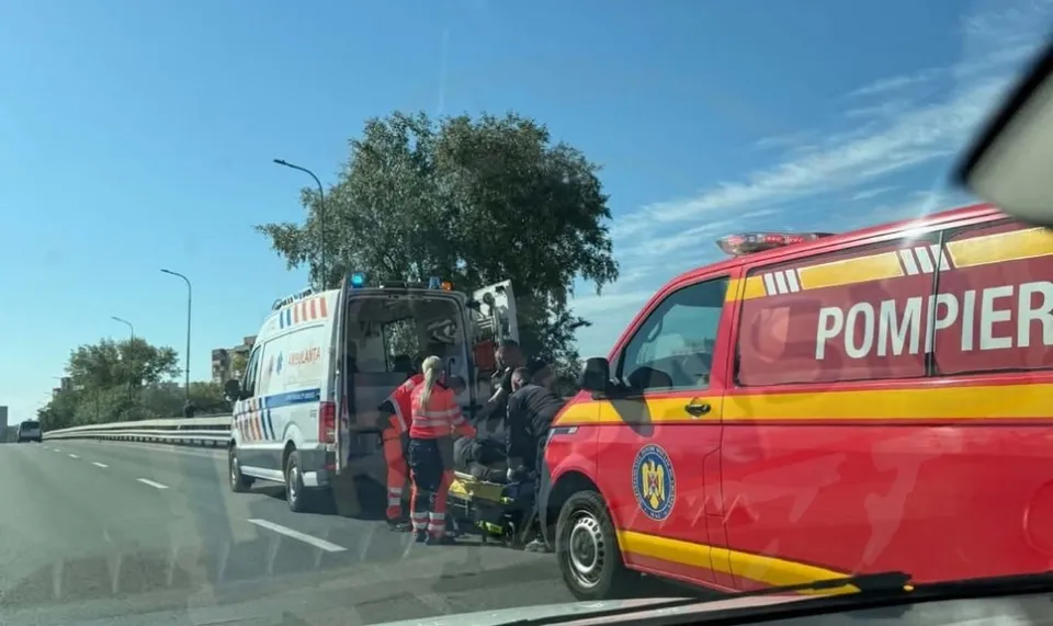 Două vehicule de urgență, una albă și una roșie, sunt oprite pe o șosea lângă un copac sub un cer senin.