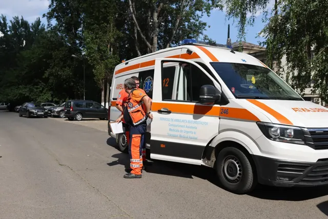 O ambulanță albă cu dungi portocalii este parcată pe un drum, lângă doi operatori medicali îmbrăcați în uniforme portocalii și albastre.