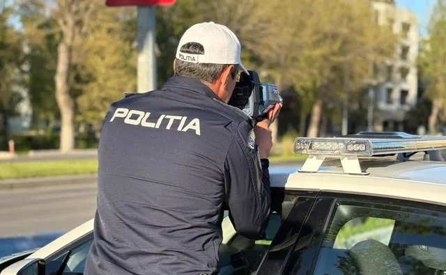 Un agent de poliție cu șapcă și uniformă neagră folosește un aparat de măsurat viteza de pe acoperișul unei mașini albe.