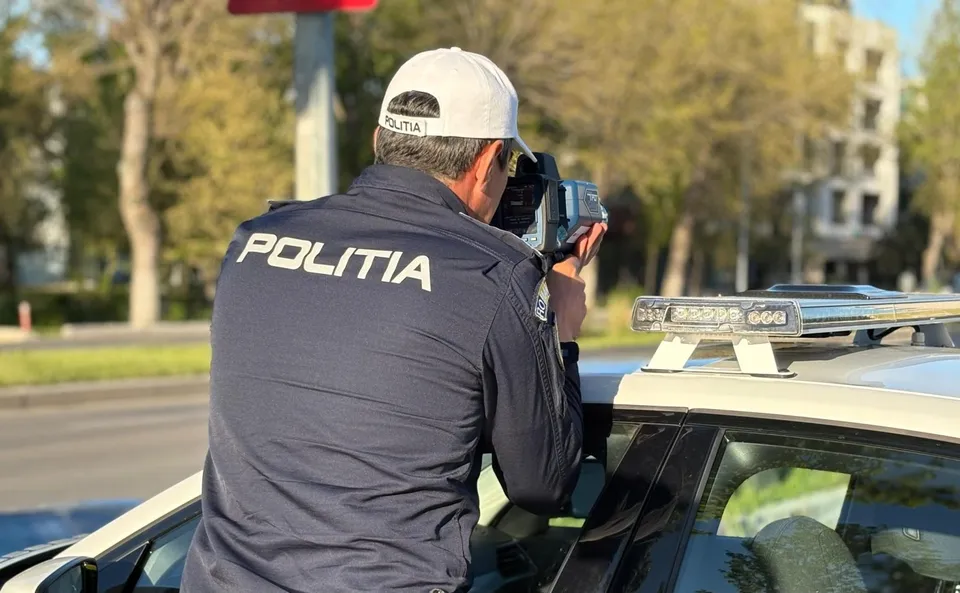 Un agent de poliție cu șapcă și uniformă neagră folosește un aparat de măsurat viteza de pe acoperișul unei mașini albe.
