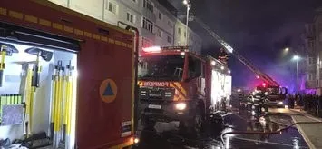 Un camion de pompieri roșu este parcat pe un drum umed lângă un bloc de locuințe, cu o scară întinsă spre acoperișul clădirii noaptea.