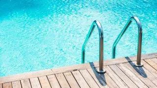 Două scări metalice de acces sunt montate pe marginea unei platforme de lemn lângă apa unei piscine azurii.