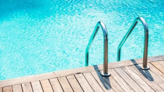 Două scări metalice de acces sunt montate pe marginea unei platforme de lemn lângă apa unei piscine azurii.