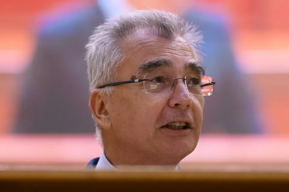 Un bărbat cu părul cărunt și ochelari cu ramă subțire privește spre lateral într-un cadru de prim-plan.