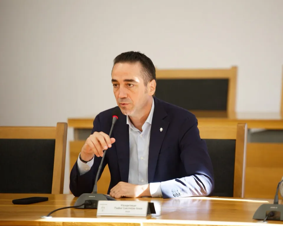 Un bărbat cu sacou închis și cămașă deschisă stă la o masă de lemn și vorbește lângă un microfon de birou.