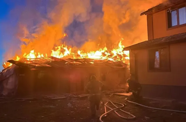 Flăcări mari și fum dens ies de pe acoperișul unei clădiri de lângă o construcție de culoare maro, în timp ce două siluete se află în prim-plan.