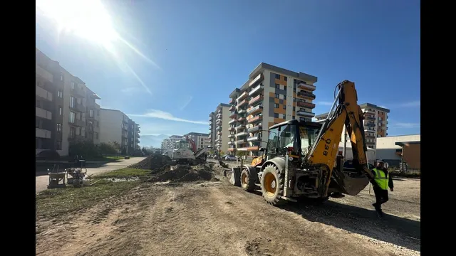 Un excavator galben stă pe un teren de pământ lângă clădiri de apartamente sub un cer senin și însorit.
