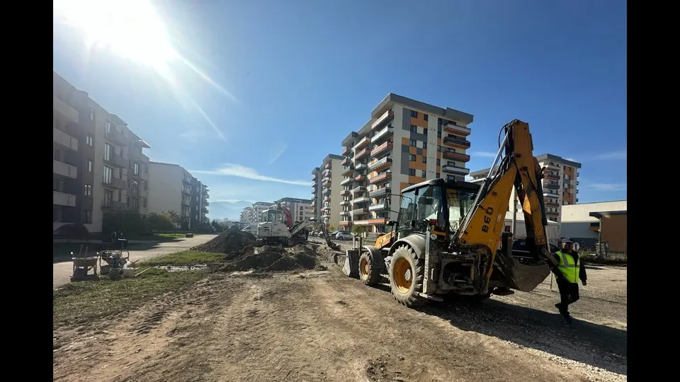 Un excavator galben stă pe un teren de pământ lângă clădiri de apartamente sub un cer senin și însorit.