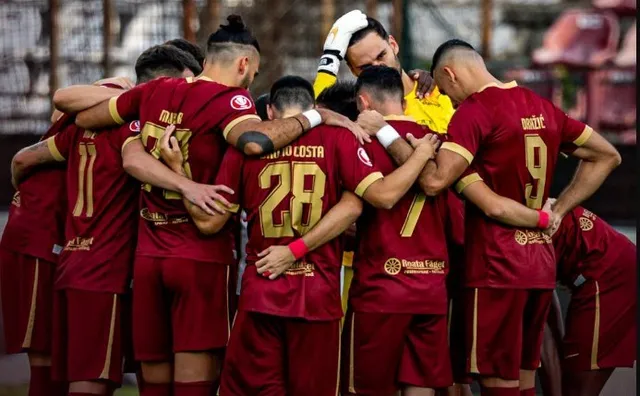 Un grup de fotbaliști în echipamente de culoare roșu închis stau într-un cerc, cu brațele pe umerii colegilor.