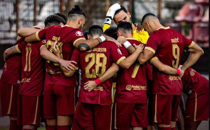 Un grup de fotbaliști în echipamente de culoare roșu închis stau într-un cerc, cu brațele pe umerii colegilor.
