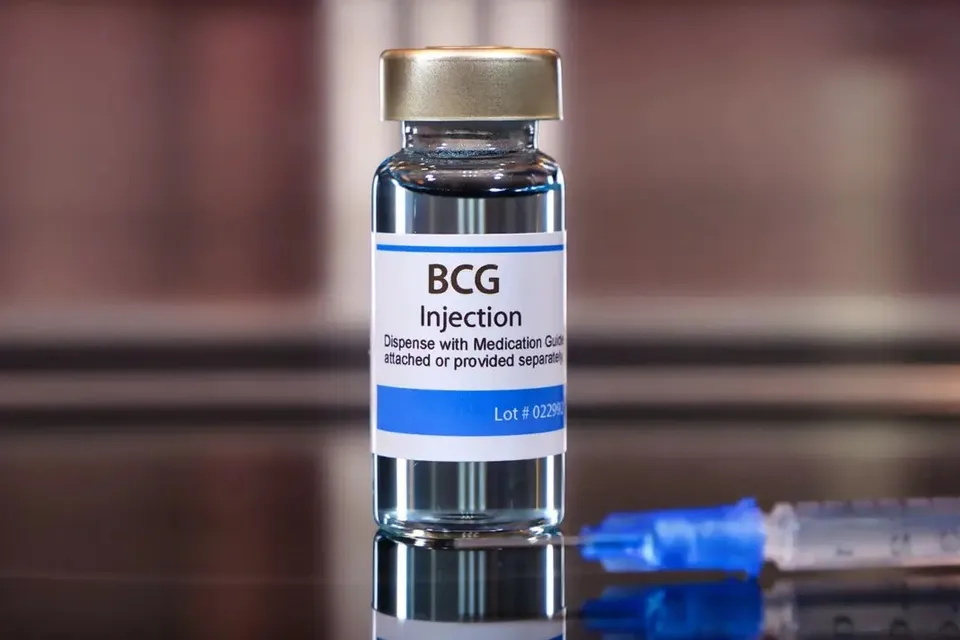Un flacon de sticlă cu etichetă albă pe care scrie BCG Injection stă pe o suprafață lucioasă lângă o seringă cu capac albastru.