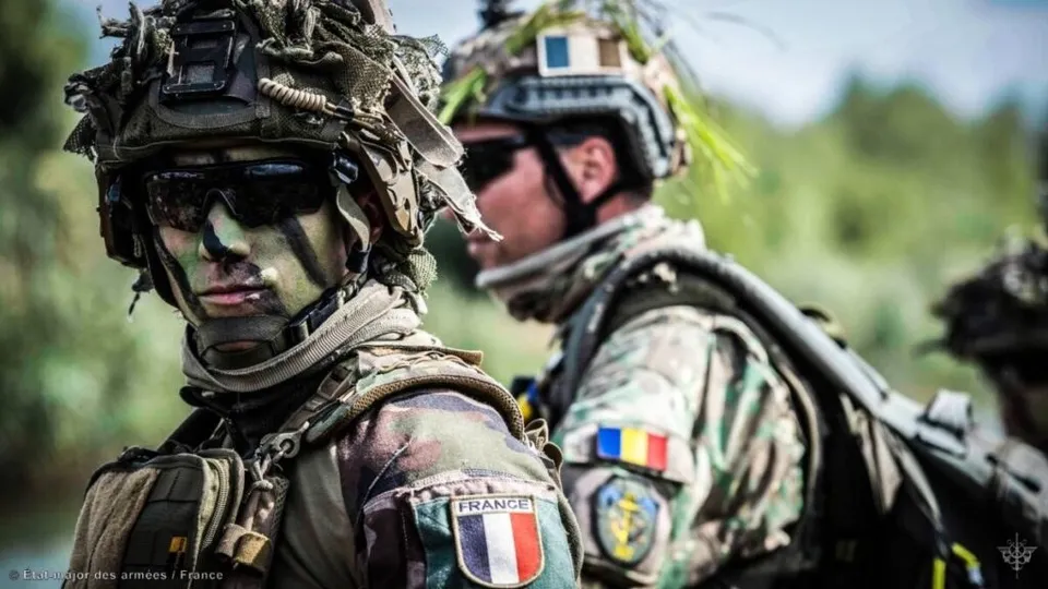Doi soldați în uniforme de camuflaj și echipament militar, purtând căști și ochelari de protecție, sunt poziționați într-un peisaj verde.