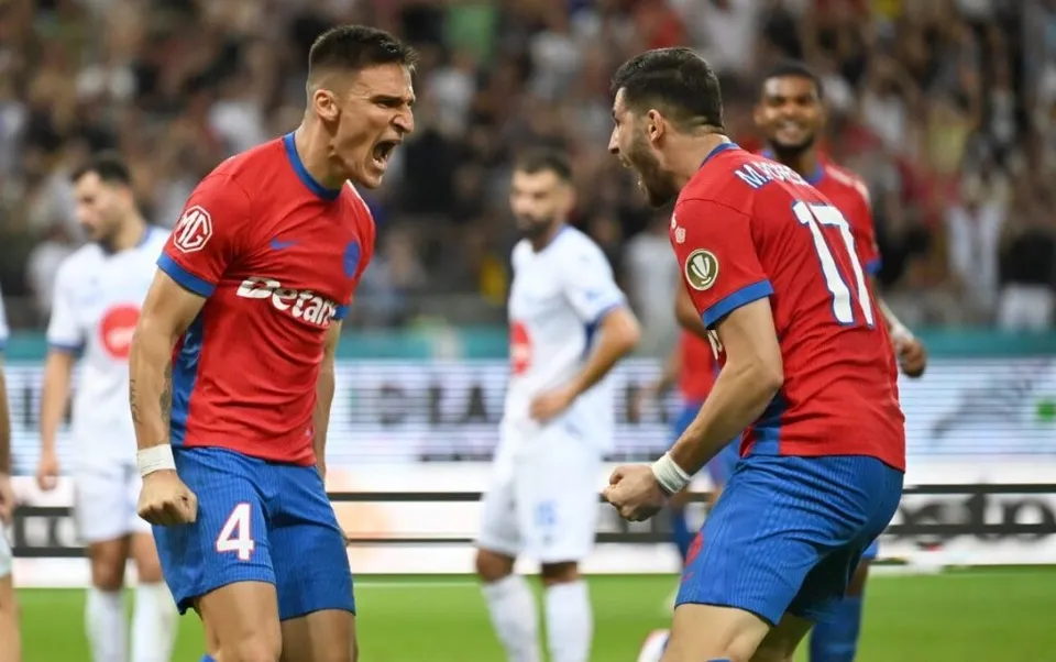 Unii jucători de fotbal în echipamente roșii și albastre sărbătoresc pe un teren de fotbal în fața unui public numeros.