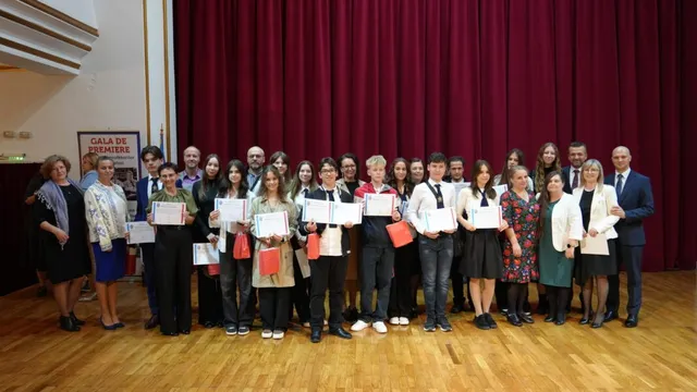 Un grup de persoane stă în rând pe un podium de lemn în fața unui paravan roșu, ținând diplome albe.