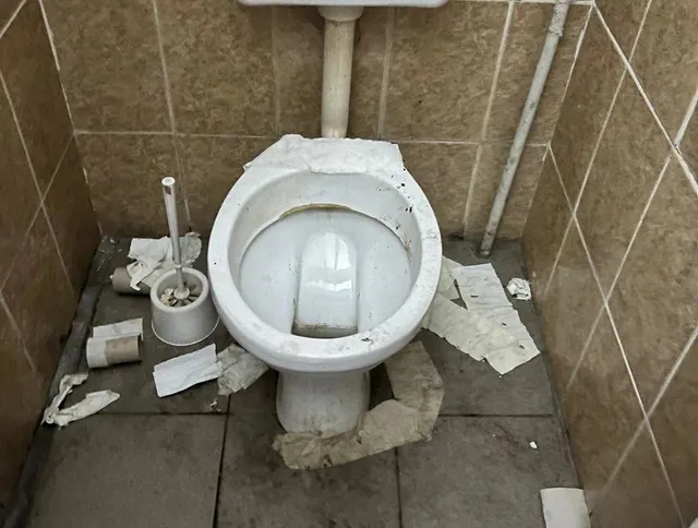 Un vas de toaletă alb este situat într-o încăpere cu plăci de faianță maro, înconjurat de resturi de hârtie pe podeaua gri.