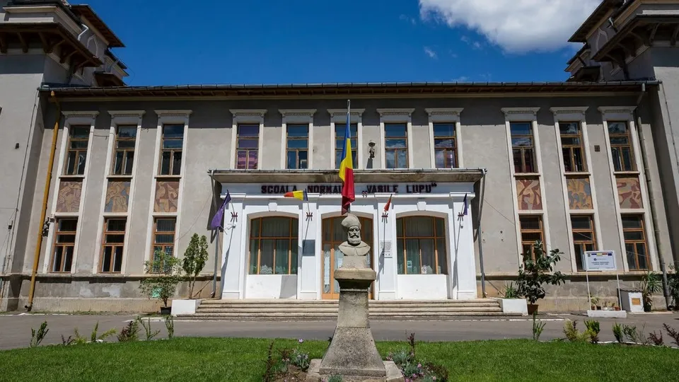 O clădire gri cu ferestre înalte, având un bust pe un soclu în prim-plan și câteva steaguri afișate la intrare.