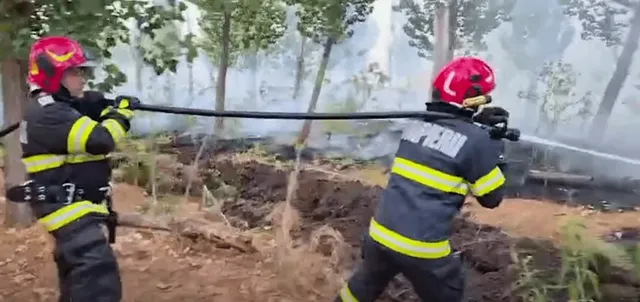 Doi pompieri în uniforme și căști roșii sting un incendiu de vegetație într-o zonă împădurită, cu fum vizibil în fundal.