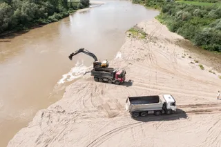 Două camioane parcate pe o zonă nisipoasă de lângă un râu, unul fiind încărcat de o excavatoare cu braț negru.