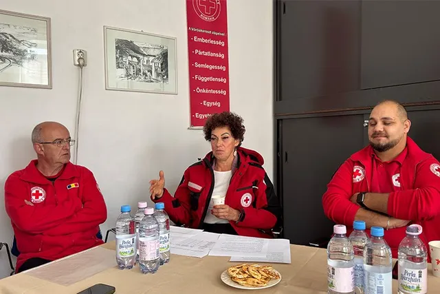 Trei persoane purtând jachete roșii cu embleme albe stau la o masă pe care se află sticle de apă și documente pe fundalul unui perete alb.