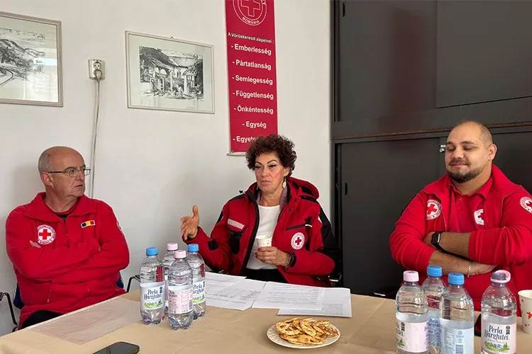 Trei persoane purtând jachete roșii cu embleme albe stau la o masă pe care se află sticle de apă și documente pe fundalul unui perete alb.