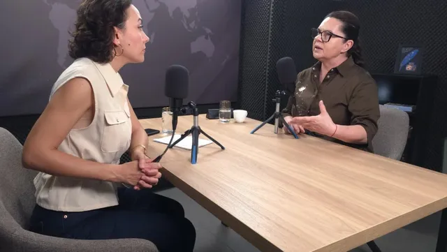 Două femei stau la o masă de lemn în fața unor microfoane, într-un studio cu fundal întunecat și un glob terestru pe perete.