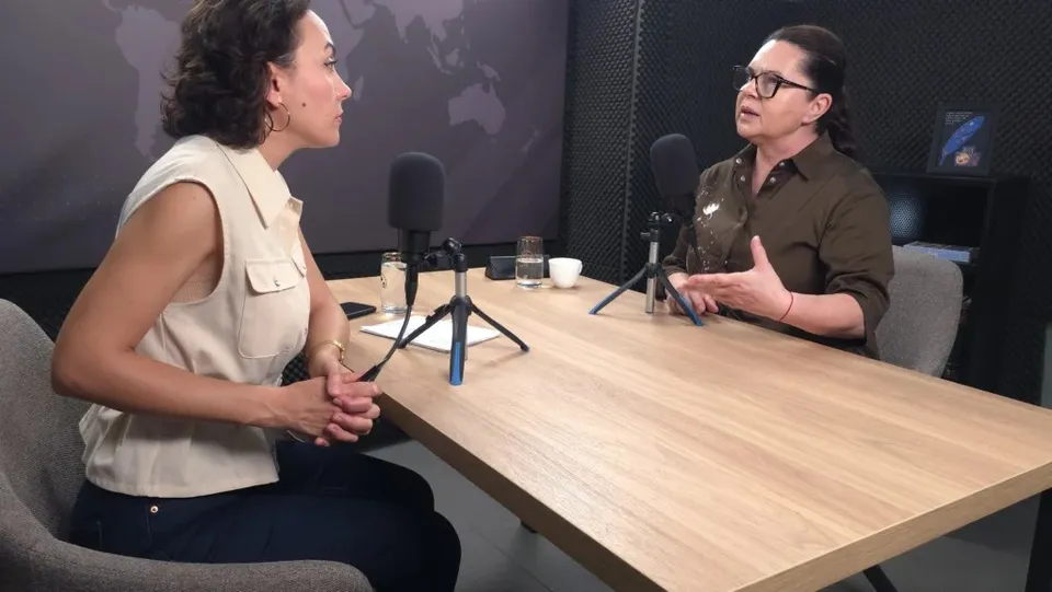 Două femei stau la o masă de lemn în fața unor microfoane, într-un studio cu fundal întunecat și un glob terestru pe perete.