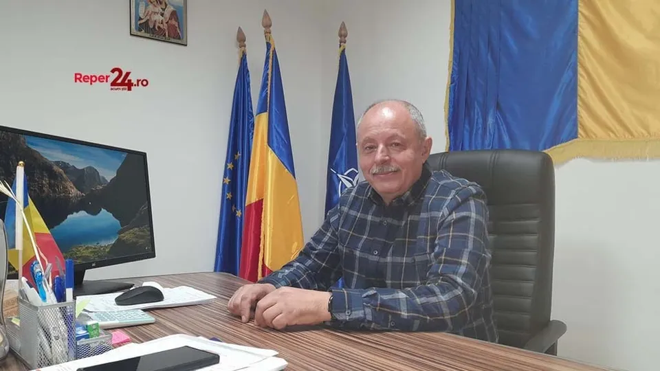 Un bărbat cu cămașă în croit este așezat la un birou lângă un monitor, trei steaguri și un scaun de piele neagră.