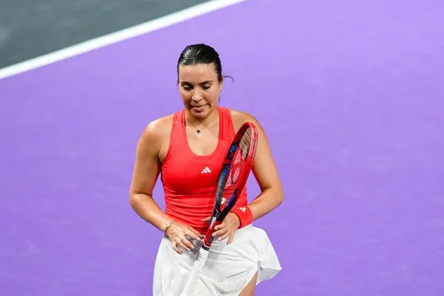 O femeie cu părul negru, purtând un tricou roșu și o fustă albă, ține o rachetă de tenis pe un teren mov.