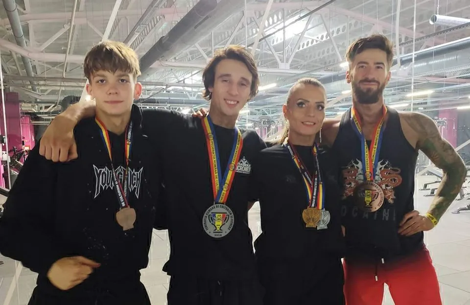 Patru persoane cu medalii la gât stau în rând într-o sală de sport cu structură metalică la tavan.