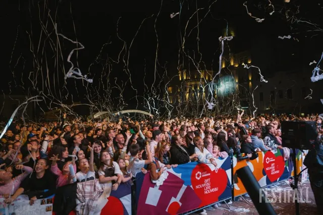 O mulțime de oameni privește în sus spre cerul de noapte, în timp ce benzi albe de confetti plutesc deasupra lor.