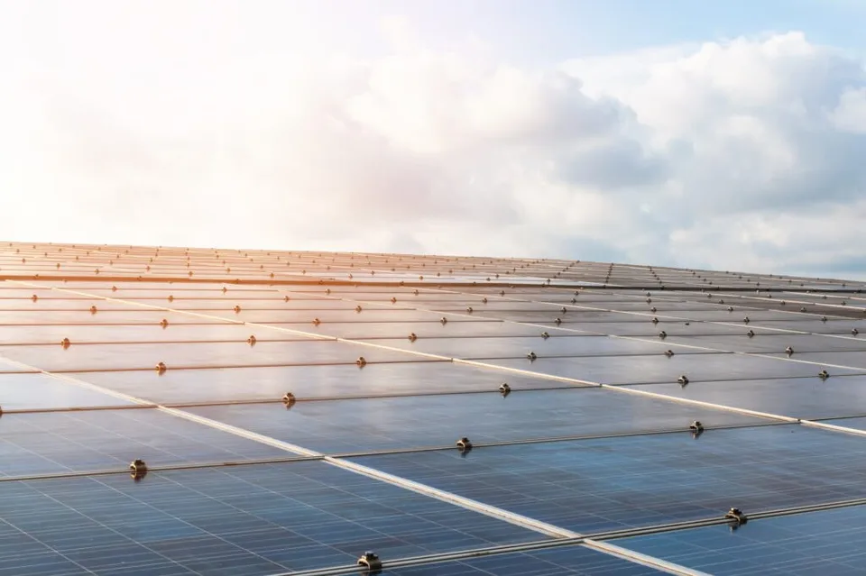 Rânduri de panouri fotovoltaice sunt instalate pe un teren sub un cer înconjurat de nori și lumină solară.