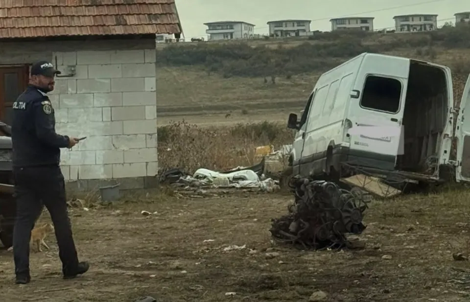 Un bărbat în uniformă întunecată stă lângă un clădire mică, privind spre un van alb cu portierele deschise pe un teren cu resturi.