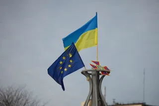 Două steaguri, unul albastru cu stele galbene și unul cu benzi orizontale albastre și galbene, sunt ridicate pe un stâlp sub un cer înnorat.