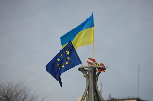 Două steaguri, unul albastru cu stele galbene și unul cu benzi orizontale albastre și galbene, sunt ridicate pe un stâlp sub un cer înnorat.