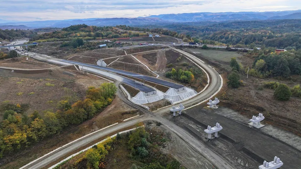 O vedere aeriană prezintă un șantier de construcții cu drumuri de acces, pământ excavat și structuri de beton albe pe un teren accidentat.