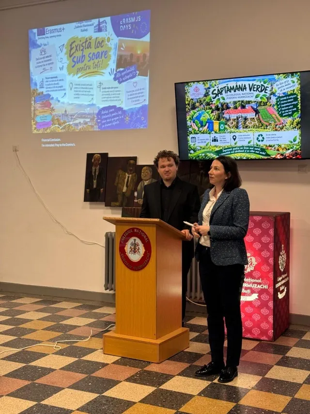 Un bărbat și o femeie stau lângă un podium de lemn într-o sală cu ecran de proiecție și panouri informative pe perete.